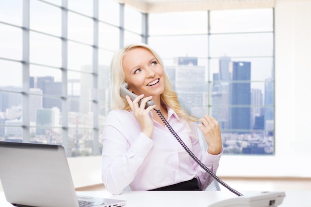 business, communication, people and technology concept - smiling businesswoman or secretary with laptop computer calling on telephone over office room with city view window background