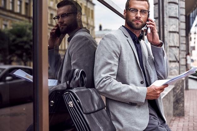 Business communications. Young businessman standing leaning on wall on the city street talking on smartphone with partner reading documents looking aside pensive close-up