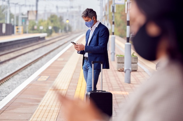 전염병 동안 PPE 안면 마스크를 착용한 휴대전화로 철도 플랫폼에서 비즈니스 통근