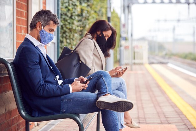 Photo business commuters sitting on railway platform with mobile phones wearing face masks during pandemic