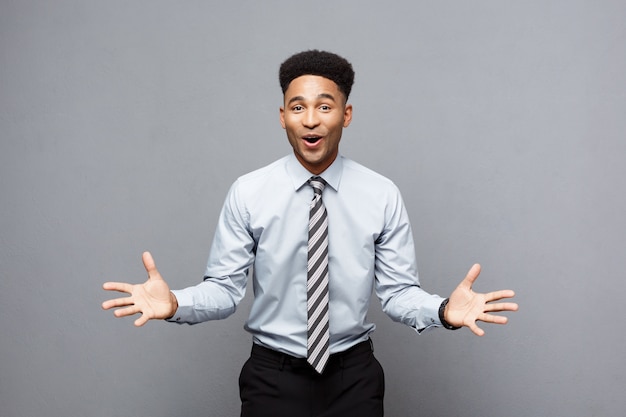 Business Concept - Confident cheerful young African American showing hands in front of him with surprising expression over grey background.