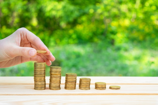 Business concept. a hand lays a coin on a column of coins on a\
wooden table with a green bokeh background. success and business\
growth. the accumulation of money. copy space.