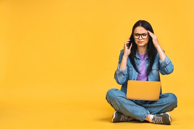 Business concept Portrait of depressed angry woman in casual sitting on floor in lotus pose and holding laptop isolated over yellow background Using phone