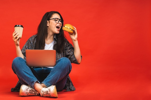 Il concetto di business ritratto di donna bruna felice in casual seduta sul pavimento in posa di loto e tenendo laptop isolato su sfondo rosso bere caffè con hamburger