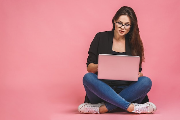 Business concept. Portrait of happy woman in casual sitting on floor in lotus pose and holding laptop isolated over pink background