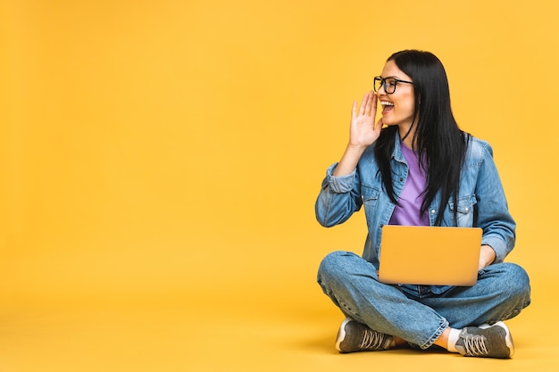 Business concept Portrait of happy young woman in casual sitting on floor in lotus pose and holding laptop isolated over yellow background Great news