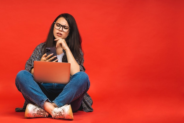 Business concept Portrait of serious brunette woman in casual sitting on floor in lotus pose and holding laptop isolated over red background Using mobile phone