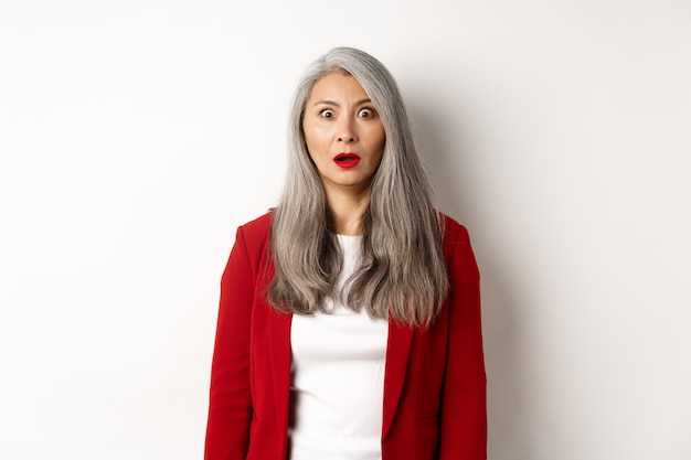 Фото Бизнес-концепция. шокированная старшая женщина-работодатель безмолвно смотрит в камеру, стоя в красном пиджаке и макияже на белом фоне