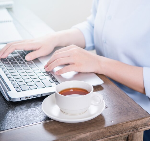 Business concept. Woman in blue shirt typing on computer with coffee on office table, backlighting, sun glare effect, close up, side view, copy space