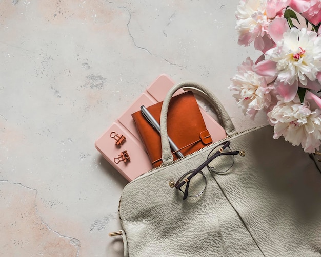 Фото Бизнес-концепция женская кожаная белая сумка с планировщиком и планшетом с пионскими цветами на светлом бетонном фоне, вид сверху