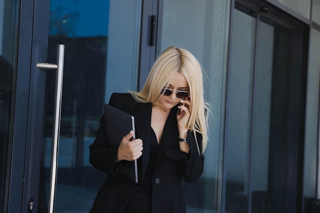 Business conceptA young woman in a suit is talking on the phone and holding a laptop in her hands on the street