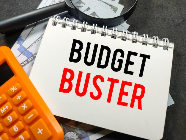 Бизнес-концепцияText BUDGET BUSTER на ноутбуке с калькулятором, увеличительным стеклом и долларовой банкнотой на черном фоне