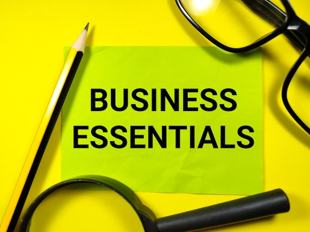 노란색 배경에 연필안경과 돋보기가 있는 종이 노트에 비즈니스 개념텍스트 BUSINESS ESSENTIALS