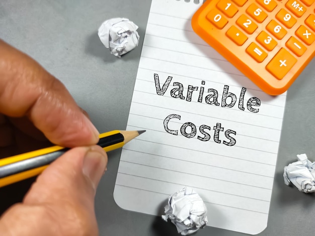 Бизнес-концепцияТекстовая переменная стоимость, написанная на блокноте с рукой, держащей карандашную бумагу и калькулятор на сером фоне