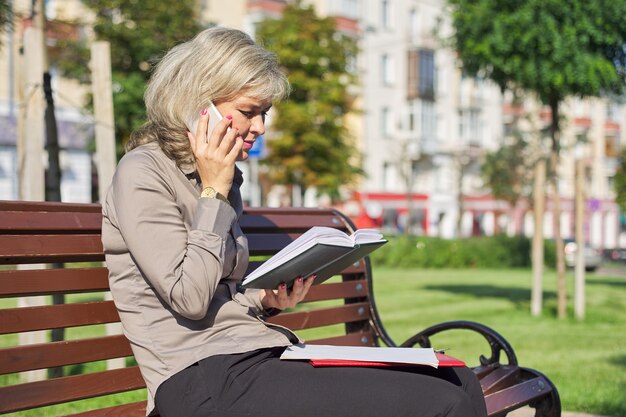 Business confident mature woman talking on cell phone, successful female in city sitting on bench, looking at business notebook