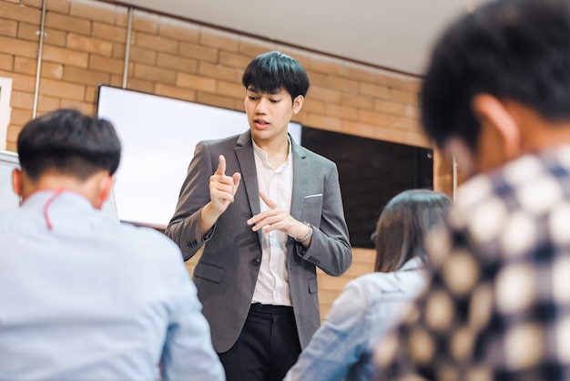 Business cooperation : young asian male coach or speaker make\
flip chart presentation to diverse businesspeople at meeting in\
office. male tutor or trainer present project to diverse\
colleagues.