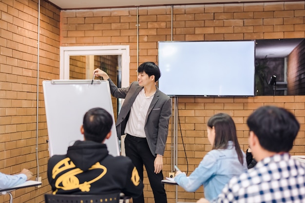 Business cooperation : young asian male coach or speaker make\
flip chart presentation to diverse businesspeople at meeting in\
office. male tutor or trainer present project to diverse\
colleagues.