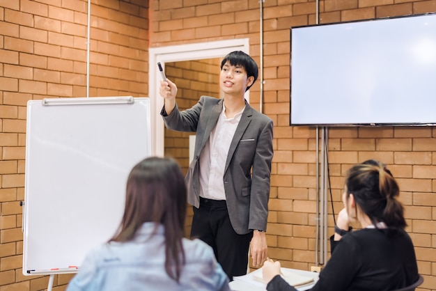 Business cooperation : young asian male coach or speaker make\
flip chart presentation to diverse businesspeople at meeting in\
office. male tutor or trainer present project to diverse\
colleagues.