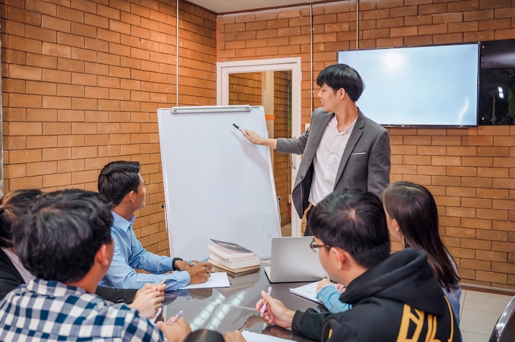 Photo ビジネス協力：若いアジア人男性のコーチまたはスピーカーが、オフィスでの会議で多様なビジネスマンにフリップチャートプレゼンテーションを行います。男性の家庭教師またはトレーナーがさまざまな同僚にプロジェクトを提示します。