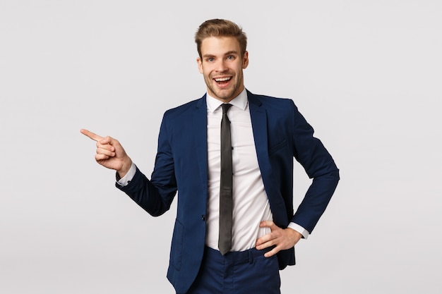 Business, corporate and entrepreneur concept. Handsome cheerful blond bearded businessman in suit, talking to coworkers, smiling happy and relieved, finish office meeting, pointing left