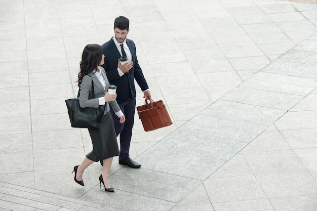 Business couple walking outdoors, talking and drinking take out coffee