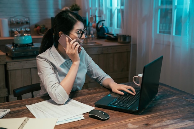 Business deadline and technology concept. asian japanese woman\
employee talking on smartphone and using laptop computer working at\
night home kitchen. female worker communicate on cellphone\
working