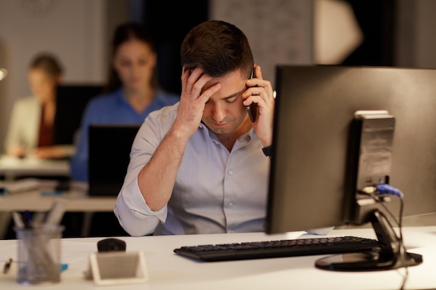 Business deadline e concept tecnologico uomo d'affari con computer che chiama su sartphone in ufficio notturno