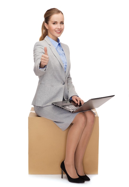 business and delivery service concept - smiling woman sitting on cardboard box with laptop computer showing thumbs up