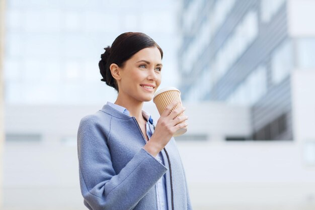 концепция бизнеса, напитков, отдыха и людей - улыбающаяся женщина пьет кофе над офисным зданием в городе
