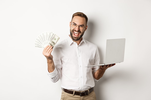 Business and e-commerce. Excited businessman holding money dollars and laptop, working online, standing over white background.