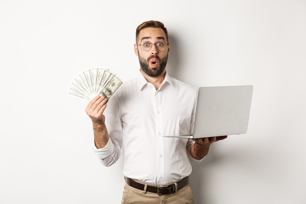 Business and e-commerce. Man looking amazed with money income, working online, using laptop, standing over white background.