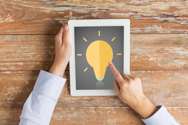 business, education, inspiration, people and technology concept - close up of hands with lighting bulb icon on tablet pc computer screen and coffee cup at table