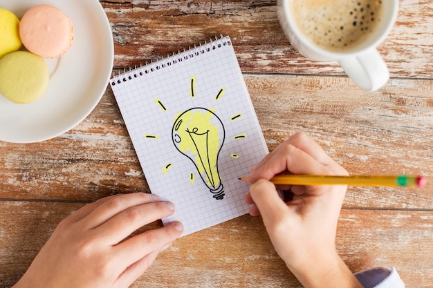 business, education and people concept - close up of female hands with pencil, coffee and cookies drawing lighting bulb to notebook on table