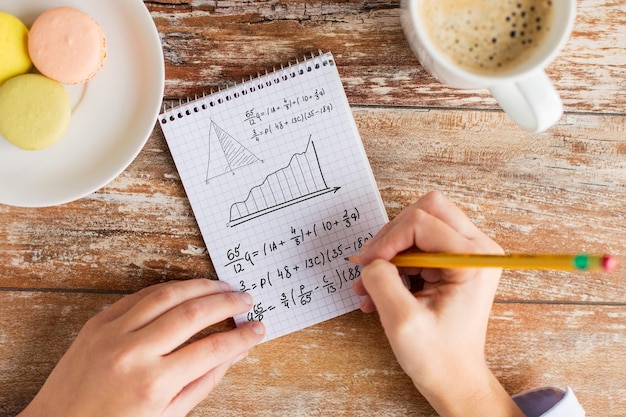 Concetto di affari, istruzione e persone - primo piano delle mani femminili con matita, caffè e biscotti per risolvere compiti o scrivere equazioni matematiche sul taccuino