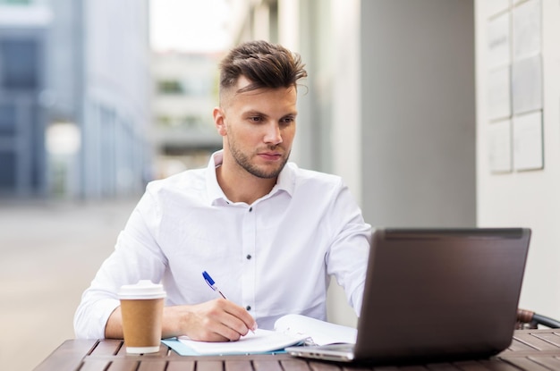 business, education, technology and people concept - young man with laptop computer, documents and coffee cup at city street cafe