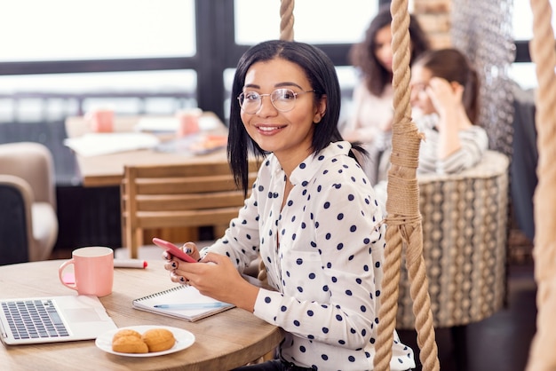 Business email. Gay kind charming businesswoman gazing at camera while carrying phone and grinning