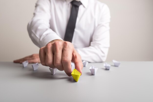 Business executive sitting at his desk pushing forward a yellow colored paper boat to advance and stand out from the crowd of other white paper boats