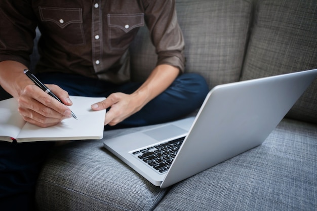 business executive working on laptop and writing on a notebook while sitting sofa at home office