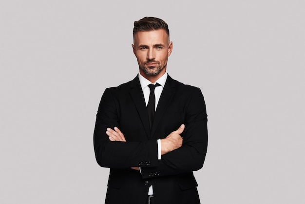 Business expert. Handsome young man in full suit keeping arms crossed and looking at camera while standing against grey background