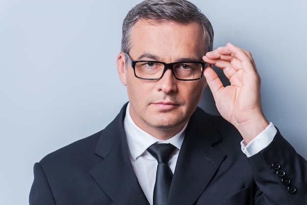 Business expert. Portrait of confident mature man in formalwear adjusting his eyeglasses and looking at camera  while standing against grey background