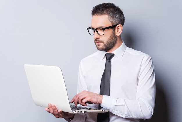 Business expert at work. Confident mature man in shirt and tie working on laptop while standing against grey background