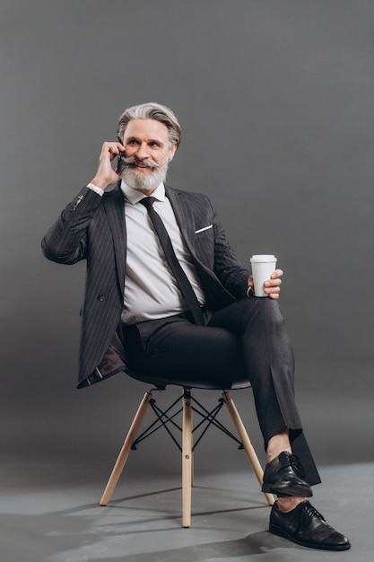 Business and fashionable bearded mature man in a gray suit sitting and taking by phone on the grey wall.