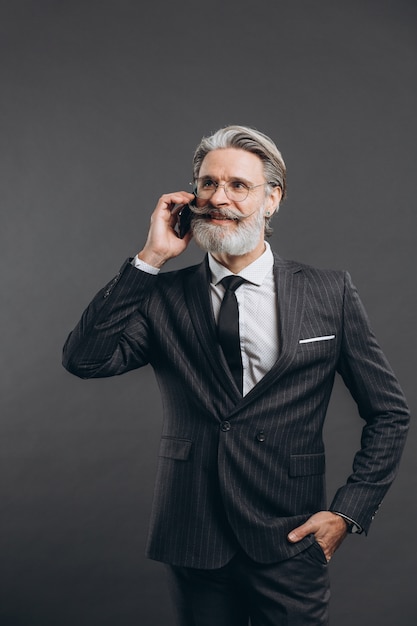 Business and fashionable bearded mature man in a gray suit smiling and talking by phone on the grey wall.