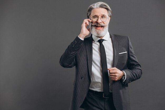 Business and fashionable bearded mature man in a gray suit smiling and talking by phone on the grey wall.