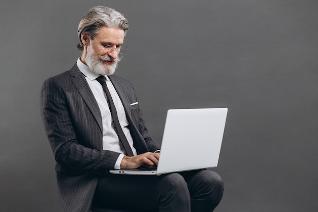 Business and fashionable bearded mature man in a gray suit smiling and using laptop on the grey wall.