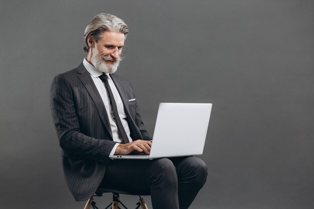 Business and fashionable bearded mature man in a gray suit smiling and using laptop on the grey wall.