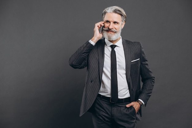 Business and fashionable bearded mature man in a gray suit talking by phone and smile on the grey wall.