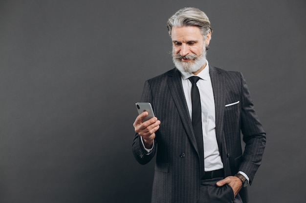 Business and fashionable bearded mature man in a gray suit talking on the phone and smile on the grey wall.