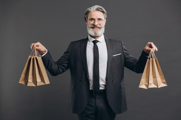 Foto uomo maturo barbuto alla moda e di affari in un vestito grigio con i pacchetti del mestiere e fare acquisti sulla parete grigia.