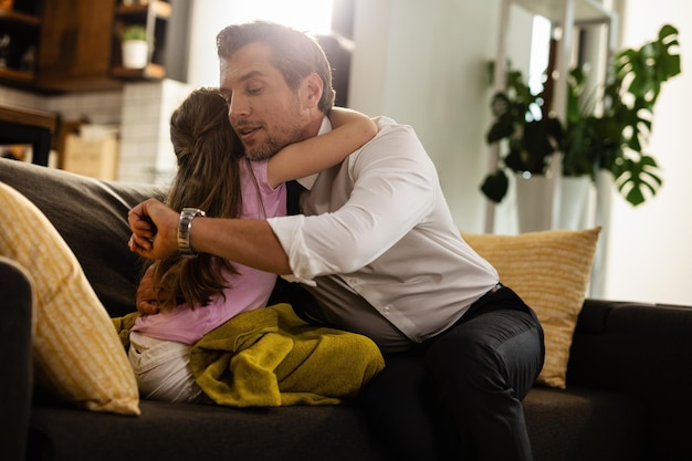 Business father checking the time on wristwatch while being embraced by his small daughter at home